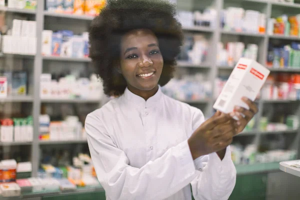 매력적 인 젊은 아프리카 약사에게 흰 상자에 새로운 약을 주입하고 카메라를 응시하면서 유리 뒤에 현대 약국에서 있는 동안 — 스톡 사진
