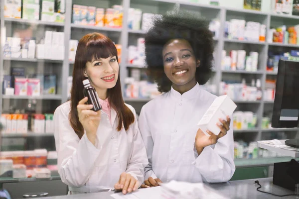 Des pharmaciennes africaines et caucasiennes souriantes posent près de la table avec un noeud de cashbow à l'apothicaire, montrant des médicaments à la caméra. Bienvenue à la pharmacie — Photo