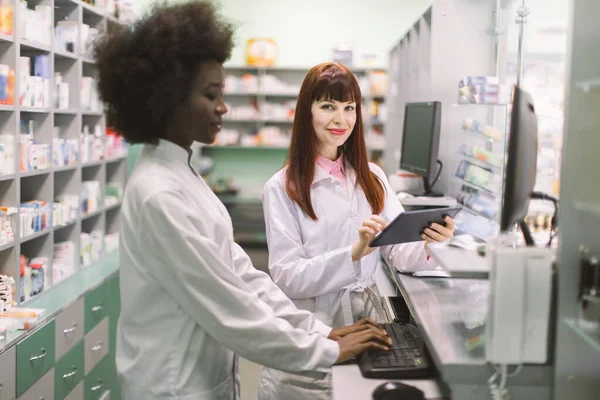 Dvě mladé veselé lékárnice, které spolu pracují v lékárně. Afričanka píše na počítači, zatímco její bělošská kolegyně používá tablet. Zaměření na bělošku — Stock fotografie