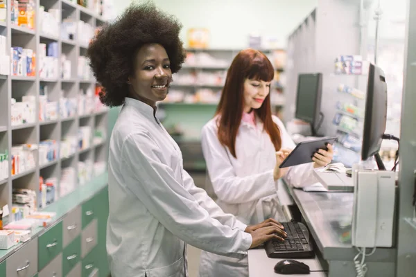 Deux jeunes pharmaciennes joyeux qui travaillent ensemble à la pharmacie. Femme africaine tapant sur ordinateur tandis que son collègue caucasien utilisant tablette. Focus sur la femme africaine souriante — Photo