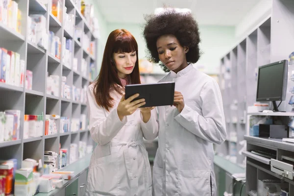 Dwie młode, pewne siebie kobiety farmaceutki wielorasowe, afrykańskie i kaukaskie, w nowoczesnej aptece, patrzące na ekran tabletu, sprawdzające leki lub składające zamówienie — Zdjęcie stockowe