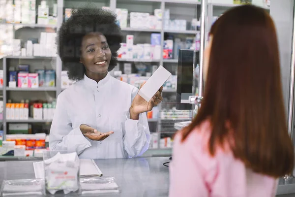Lékárna, lidé a koncept zdravotní péče. Usmívající se afrikánská lékárnice, která léčí mladou bělošku v lékárně. Pohled přes rameno pacienta — Stock fotografie