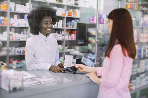 Pěkná mladá běloška, která platí lékařům kreditkou v lékárně. Usmívající se atraktivní africká žena lékárník dávkování léků pro mladé ženy v lékárně — Stock fotografie