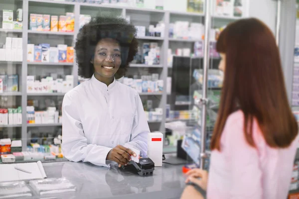 Kaukaska klientka płacąca w aptece. Afrykanka aptekarka wydająca lek, posiadająca terminal i kartę kredytową pacjentki płacącej za leki — Zdjęcie stockowe