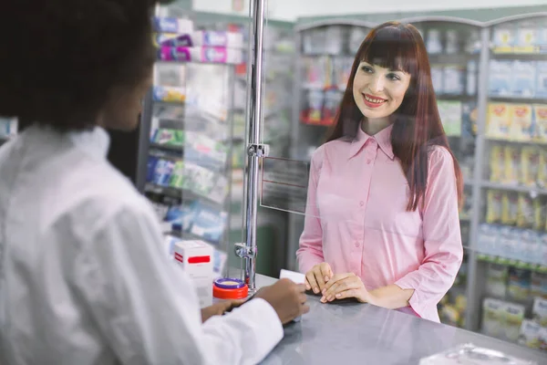 Csinos, fiatal kaukázusi nő, aki hitelkártyás gyógyszereket fizet a modern gyógyszertárban. Az afrikai női gyógyszerész gyógyszerkiadásának áttekintése fiatal nők számára a gyógyszertárban — Stock Fotó