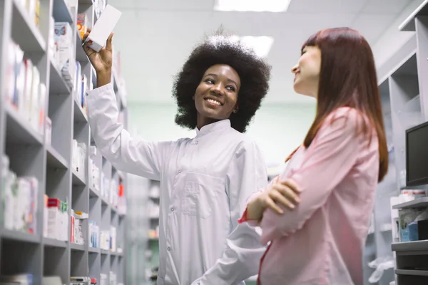 Młoda, pogodna czarna farmaceutka rekomenduje lek bezpieczeństwa dla młodej białej klientki w nowoczesnej aptece. Koncepcja opieki farmaceutycznej — Zdjęcie stockowe