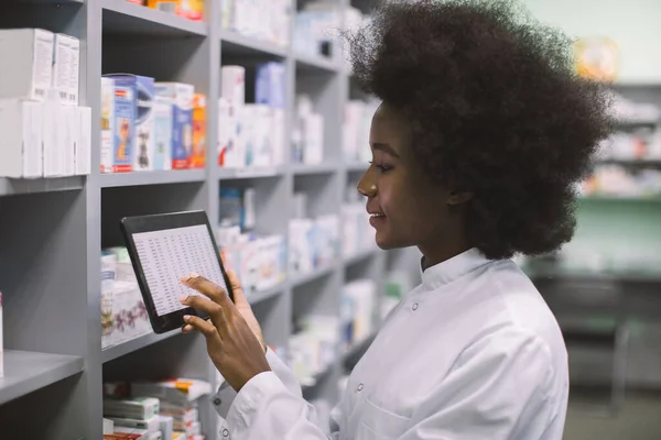 Zamknij widok z boku młodej, uśmiechniętej afrykańskiej farmaceutki korzystającej z tabletu cyfrowego podczas inwentaryzacji lub porządkującej leki w nowoczesnej aptece — Zdjęcie stockowe