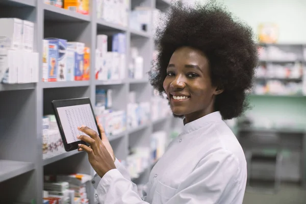 Młoda, uśmiechnięta Afroamerykanka o ciemnej skórze, farmaceutka używająca cyfrowej tabletki podczas inwentaryzacji lub porządkująca leki w nowoczesnej szpitalnej aptece. — Zdjęcie stockowe