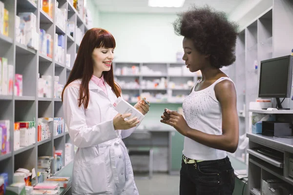 예쁘고 전문적 인 여성 약사가 젊은 아프리카 여성 환자와 이야기를 나누면서 그녀에게 두 가지 복용량의 약을 보여 주고 어떻게 사용하는지 설명 했습니다. 약물 치료 — 스톡 사진
