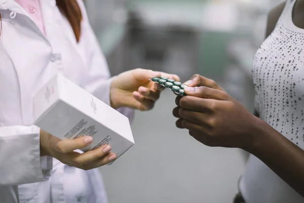 Oříznutý obrázek rukou lékárnice držící krabici s drogami a dávající puchýře s pilulkami černé ženě, dávající léky v moderní komunitní lékárně — Stock fotografie