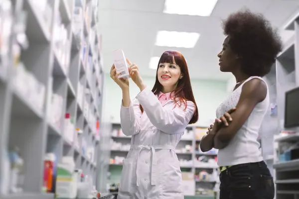 젊은 백인 여성 약사가 현대 약국에서 젊은 흑인 여성 고객에게 약을 권하고 있습니다. 여성 약사가 약을 보여 주면서 약국에 있는 고객 과상의 한다 — 스톡 사진