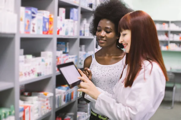 젊은 백인 여성이 웃으면서 디지탈 태블릿을 들고 있고 약국에 있는 젊은 흑인 여성에게 처방 된 약을 사용하는 방법을 주문하고 설명하는 모습 — 스톡 사진
