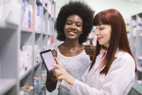 Szczęśliwy młody afrykański klient kobieta jest zadowolony z zalecanych leków w aptece, stojąc w pobliżu kobiety farmaceuta. Piękny farmaceuta patrząc na tabletkę i czytając e-receptę — Zdjęcie stockowe