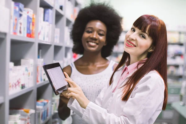 Experienced professional woman pharmacist holds tablet and consults African female customer in modern pharmacy. Pharmacist and patient at modern drugstore — Stock Photo, Image