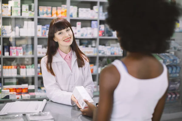 긍정적 인 젊은 백인 여성 약사가 현대 약국의 카 쉬 데스크에 서서 아프리카 여성 고객에게 약을 제공하고 있습니다. 환자의 어깨 너머를 바라봄 — 스톡 사진