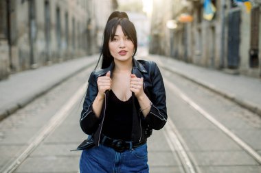 Güzel, genç, Asyalı, melez, şık, siyah deri ceket ve kot giyen, tramvay yolunda duran ve şehirde yürüyüşün tadını çıkaran bir model.