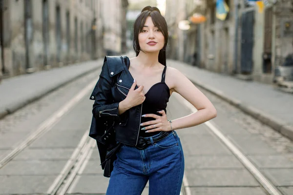 Atraktivní moderní brunetka Asijka, na sobě džíny džíny, černý top a kožené sako na rameni, pózuje na kameru se sexy tváří a úsměvem, stojí na ulici — Stock fotografie