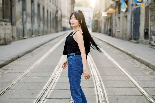 Mladá krásná asijská dívka s culíkem černé vlasy, na sobě ležérní džíny a černý top, otáčení na kameru a pózování při procházce ve městě za slunečného dne. Venkovní módní portrét — Stock fotografie