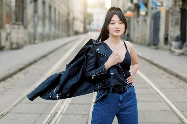 Mulher asiática corrida muito mista com longos cabelos escuros vestindo jeans e top preto, tirando seu casaco de couro preto e posando para câmera na rua da cidade. Estilo de vida urbano e moda — Fotografia de Stock