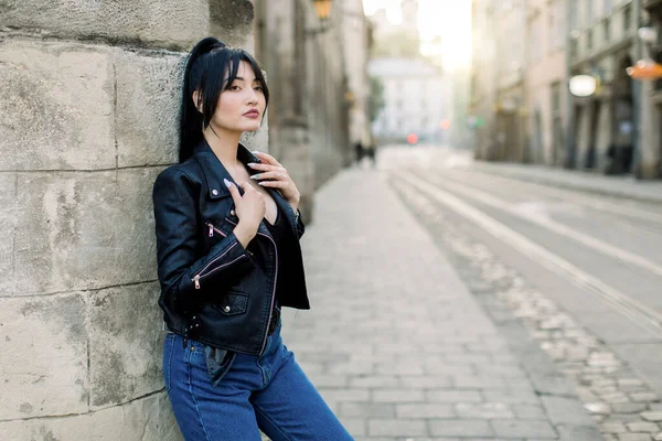 Portrét krásné brunetky Asijské módní model ve stylové kožené bundě a džínách džíny, pózování u zdi budovy. Ulice starého města a dlažba silnice na pozadí — Stock fotografie