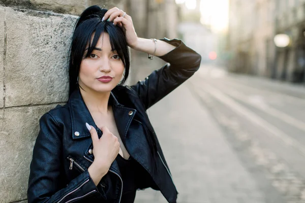 Mujer caucásica de raza mixta feliz en chaqueta de cuero negro, sonriendo mientras posaba al aire libre en la ciudad en un día soleado, apoyándose en una vieja pared de piedra. Tiro al aire libre, espacio de copia — Foto de Stock