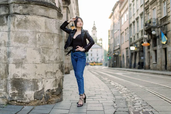 Портрет молодої жінки з брюнетки в джинсах і стильна чорна шкіряна куртка, сексуальна постава біля старої кам'яної стіни будівлі, постріл на вулиці влітку — стокове фото