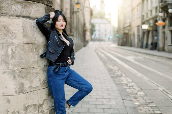 Jeune élégant modèle mixte femme brune en jeans et veste en cuir à la mode, debout dans la ville appuyé sur un mur vintage. Jolie fille mannequin dans la rue. Style de ville — Photo