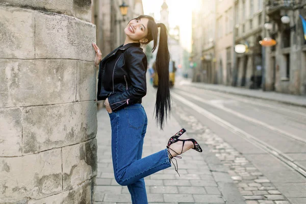 아리따운 젊은 아시아 모델인 그녀는 평범 한 검정 색 가죽 재킷과 데님과 힐을 착용하고 고대 도시 중심가에 있는 오래 된 건물 근처에 서 있다. 패션 모델, 도시 생활 방식 — 스톡 사진