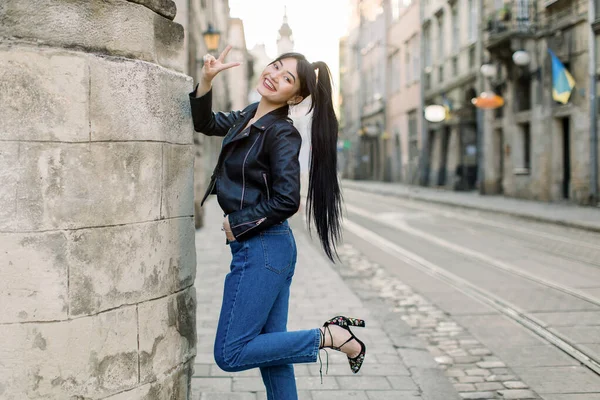 Bella giovane donna modello asiatico con i capelli coda di cavallo, in giacca di pelle nera alla moda casual e denim, e tacchi, in piedi vicino al vecchio edificio nel centro storico della città e mostrando segno di vittoria di pace — Foto Stock