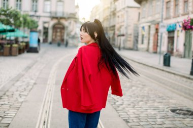 Kırmızı ceket ve kot pantolon giyen, eski Avrupa şehrinde yürüyen çekici Asyalı genç bir kadının hareketine bir şans. Moda kadını modelinin açık şehir portresi.