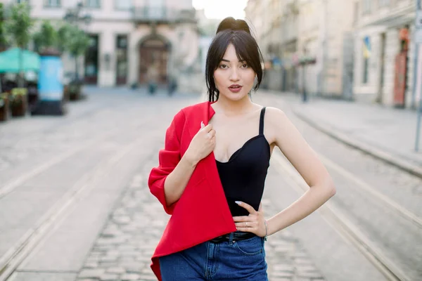 Hezká asijská sexy smyslná mladá žena, oblečená v džínách, černé svrchní a módní červené sako na rameni, pózující na kameru, zatímco stojí venku na staré městské ulici. Městský životní styl — Stock fotografie