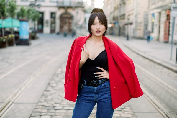 Senhora asiática bonita elegante com cabelo longo preto do rabo de cavalo, vestindo a jaqueta vermelha elegante, parte superior preta e jeans, posando à câmera na manhã do dia do verão na rua velha da cidade. Retrato urbano de rua — Fotografia de Stock