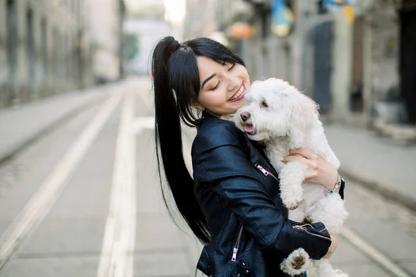 A young dark haired Asian woman, dressed in black leather jacket, stands on street in old European city street, holding and embracing white little dog. Horizontal urban shot, copy space — Stock Photo, Image