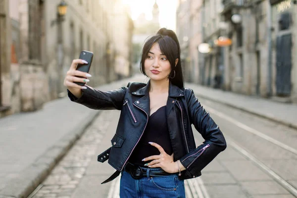 Young sexy attractive Asian woman in jeans and black leather jacket, making selfie photo on her smarphone, posing outdoors in the city, standing on the road. People, lifestyle and urban concept