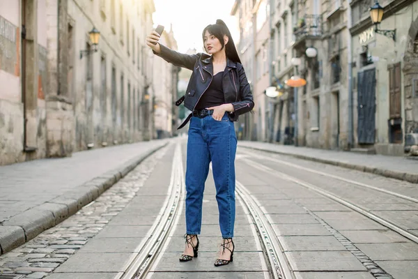 Повний горизонтальний кадр молодої веселої азіатської жінки, в модних джинсових штанях і чорній шкіряній куртці, приймаючи сельфі за мобільним телефоном, поставляючи на трамвайну дорогу на старій вулиці міста — стокове фото