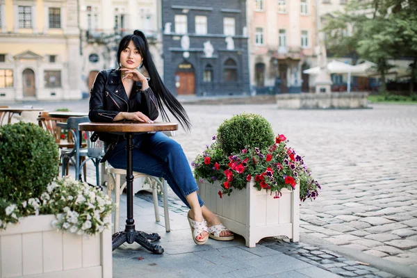 Felice donna bruna asiatica all'aperto caffè, seduto a tavola. Giovane donna elegante in jeans e giacca di pelle, a riposo all'aperto al caffè nella vecchia città europea — Foto Stock