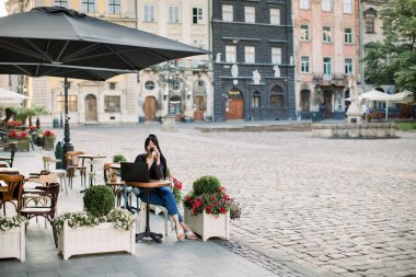 Açık şehir kafesinde dizüstü bilgisayar kullanan, masada oturan ve kahve içen genç, çekici esmer kadın. Arka planda Avrupa şehrinin eski vintage binaları