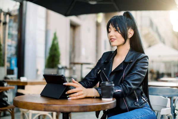 포니테일 머리의 아시아 갈색 머리 소녀가 웃고, 야외 카페에 앉아 일하는 동안 디지털 태블릿을 사용하고, 도시 카페에서 공부하고 있는 여성 대학생이 맑은 날 현대 태블릿을 사용하는 모습. — 스톡 사진