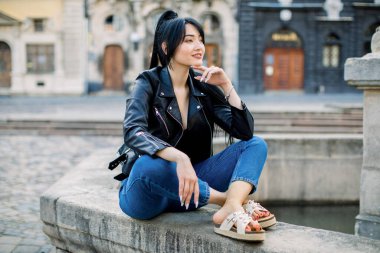 Güzel, genç, rüya gibi seksi Asyalı kadın, modaya uygun günlük giysiler içinde, eski Avrupa şehir sokağındaki taş çeşmede otururken poz veriyor ve başka tarafa bakıyor. Seyahat, yaşam tarzı, kentsel konsept