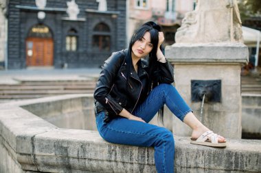 Güzel, genç, rüya gibi seksi Asyalı kadın modaya uygun günlük giysiler içinde eski Avrupa şehir sokağındaki taştan çeşmede otururken poz veriyor ve kameraya bakıyor. Seyahat, yaşam tarzı, kentsel konsept