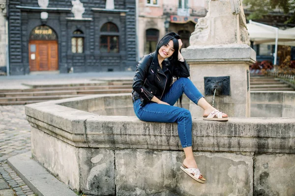 Foto all'aperto di bella hipster bruna elegante donna asiatica nella città vecchia, indossando jeans di jeans e giacca di pelle, seduto vicino alla fontana vintage Foto di moda estate, copia lo spazio. — Foto Stock