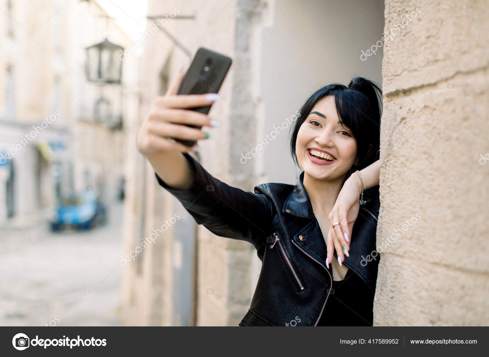 明るい若いアジアのブルネットのビジネス女性の肖像画 黒い革のジャケットを着て 屋外で彼女のスマートフォンで自撮り 古代の建物の近くの古い街の通りにポーズ ストック写真 C Sofiiashunkina Gmail Com