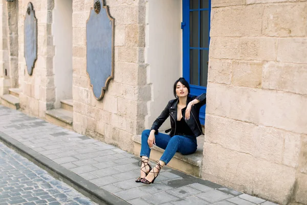 Retrato ao ar livre de jovem bela senhora asiática na moda, vestindo saltos, jeans e jaqueta de couro, sentado nas escadas do edifício antigo com porta azul, em uma rua da cidade velha. — Fotografia de Stock