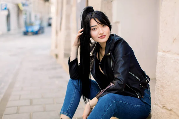 Portrét mladé krásné asijské dámy v džínových kalhotách a černé kožené bundě, sedící na kamenných schodech u staré budovy ve městě. Životní styl města. Kopírovat prostor pro text. — Stock fotografie
