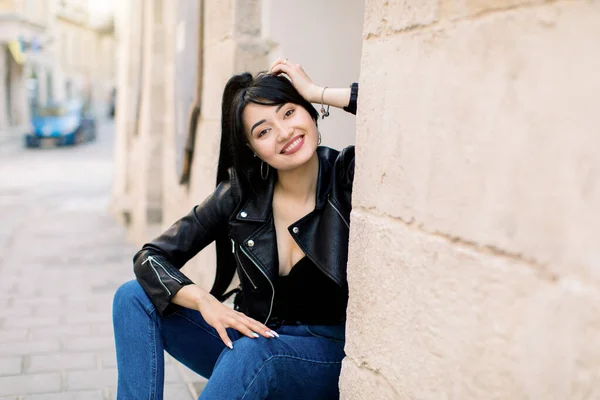 Retrato de rua de jovem bonita alegre sorrindo mulher asiática com penteado de rabo de cavalo escuro, vestindo jaqueta de couro elegante e jeans jeans, sentado perto de edifício antigo na cidade europeia — Fotografia de Stock