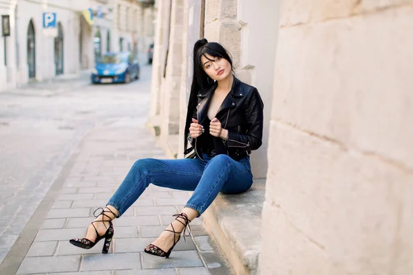 City life, people and fashion concept. Sexy young attractive Asian woman, wearing jeans, heels and black leather jacket, posing to camera outdoors in the sity, sitting on the stairs of building