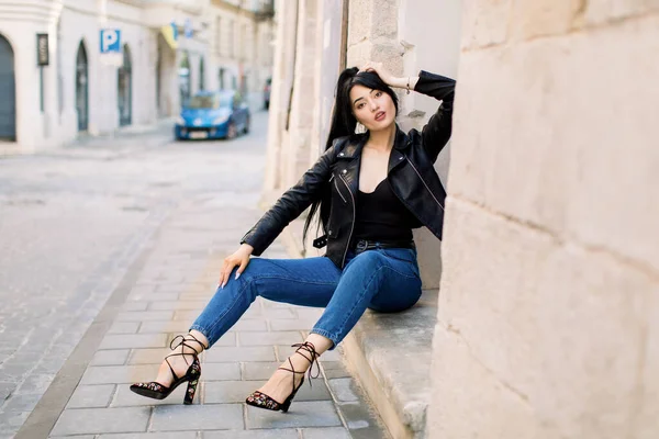Bella bruna giovane donna asiatica, indossa vestiti alla moda, jeans e giacca di pelle, seduta sulle scale di un antico edificio nella vecchia città europea. Moda città — Foto Stock