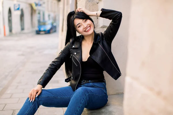 Mulher asiática jovem sexy bonita com cabelo de rabo de cavalo, vestindo roupas casuais na moda, jaqueta de couro e jeans jeans jeans, sentado ao ar livre nas escadas da cidade, sorrindo para a câmera — Fotografia de Stock