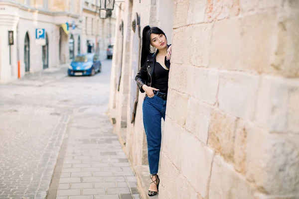 Carino attraente ragazza asiatica in giacca di pelle nera e jeans, in posa per la fotocamera all'aperto nella vecchia strada della città, nascondendo il suo corpo dietro l'antico muro dell'edificio — Foto Stock