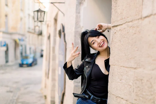 Portret van stijlvol jong Aziatisch gemengd raced vrouw, het dragen van zwart lederen jas, poseren voor de camera, staan in de buurt van oude stenen muur van het gebouw in de oude Europese stad buiten — Stockfoto